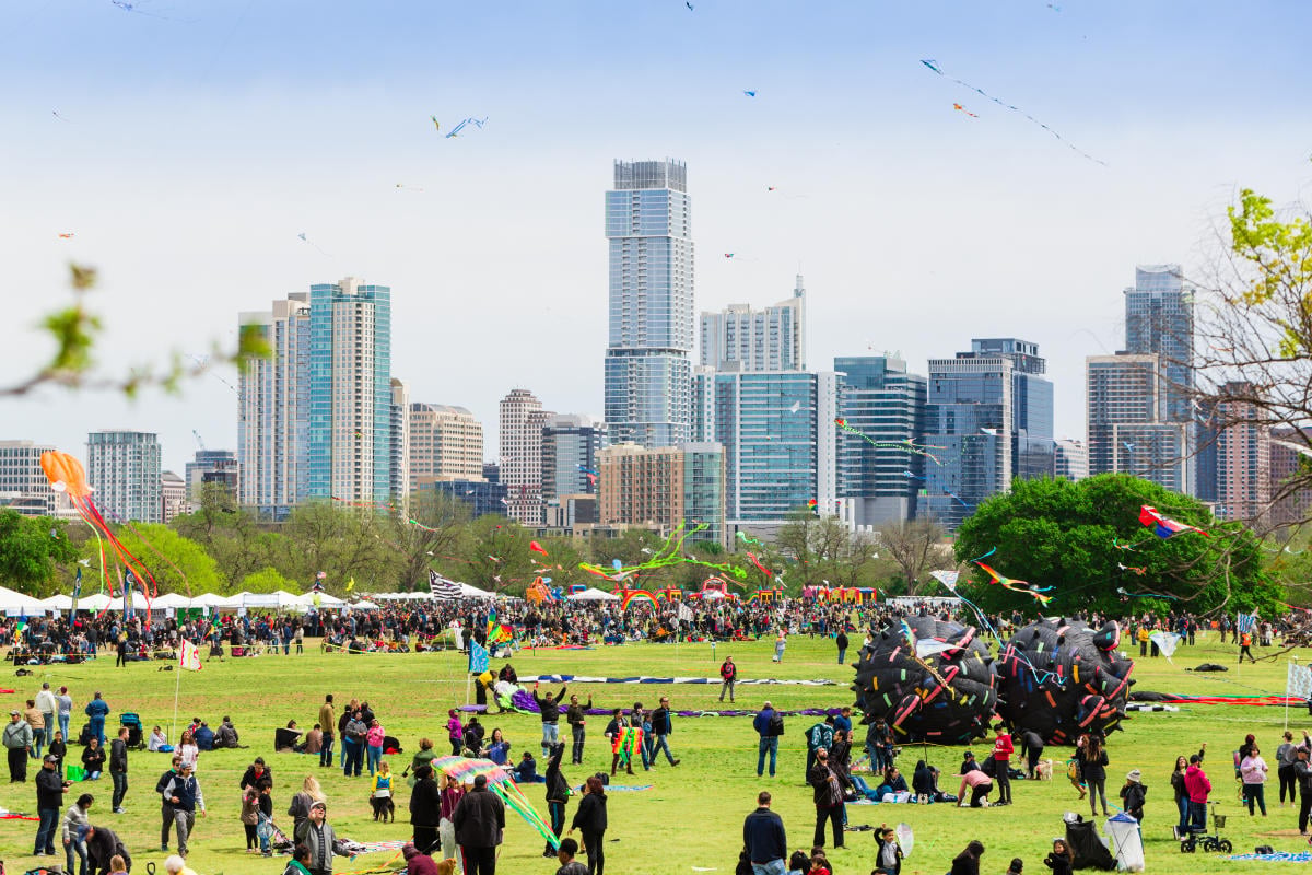 Kites and visitors in a park with Austin buildings in the distance.