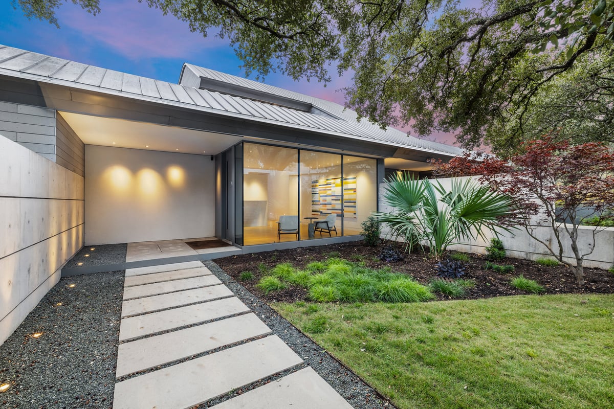 Front walk of poured concrete pavers between a concrete wall and a planted lawn, lights on inside at dusk