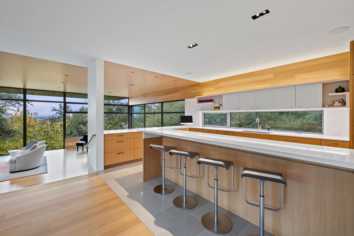 Long kitchen island with bar stools, the great room and tree canopy beyond