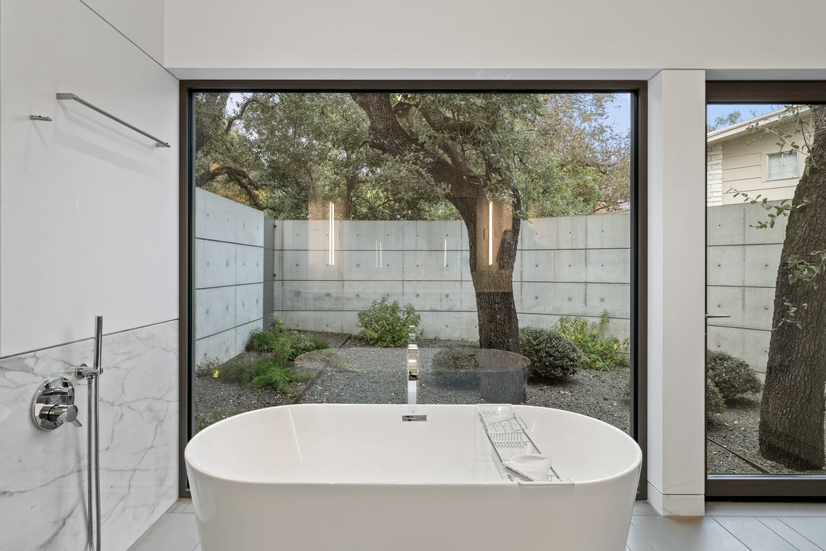 Freestanding tub facing floor-to-ceiling glass and a walled garden with a single tree