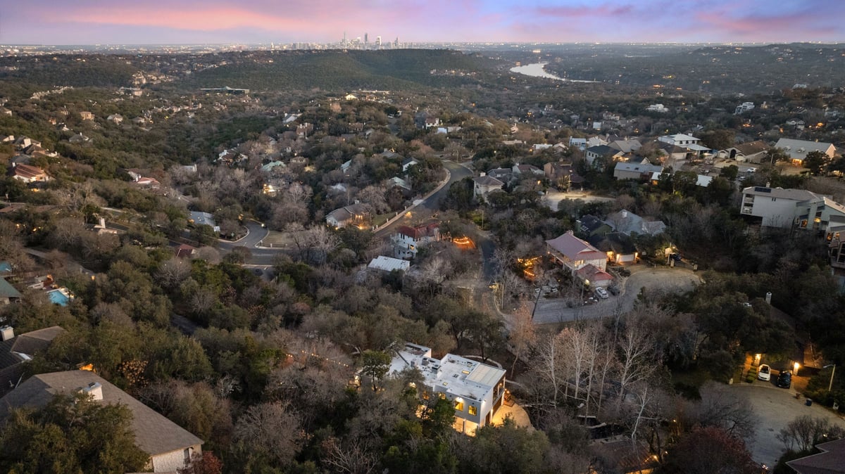 Aerial at dusk over the hillside canopy toward the Hill Country and Lake Austin