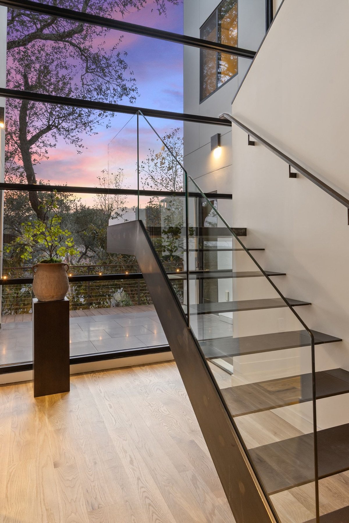 The floating glass-railed stair beside a two-story window at dusk