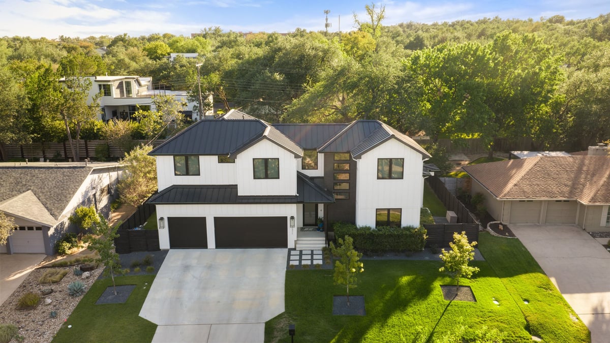 Aerial view of 7311 Running Rope from the street: a two-story white home with a dark metal roof among Northwest Hills live oaks