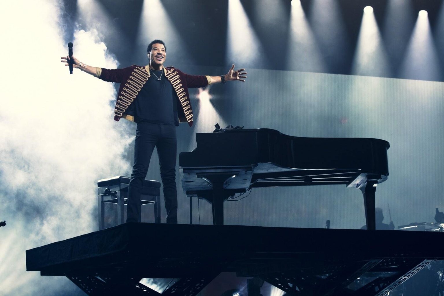 Lionel Richie performing beside a piano under stage lighting.