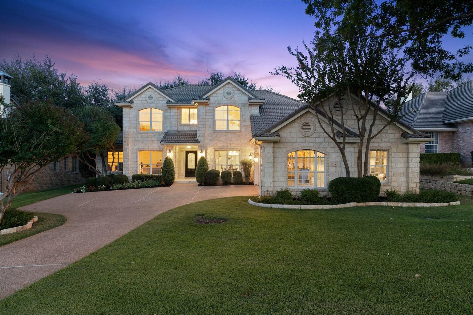 Two-story limestone home with curved drive and lawn at dusk