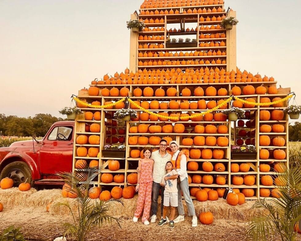 Visitors pose in front of a tall barn-shaped display stacked with pumpkins, beside a vintage red truck.