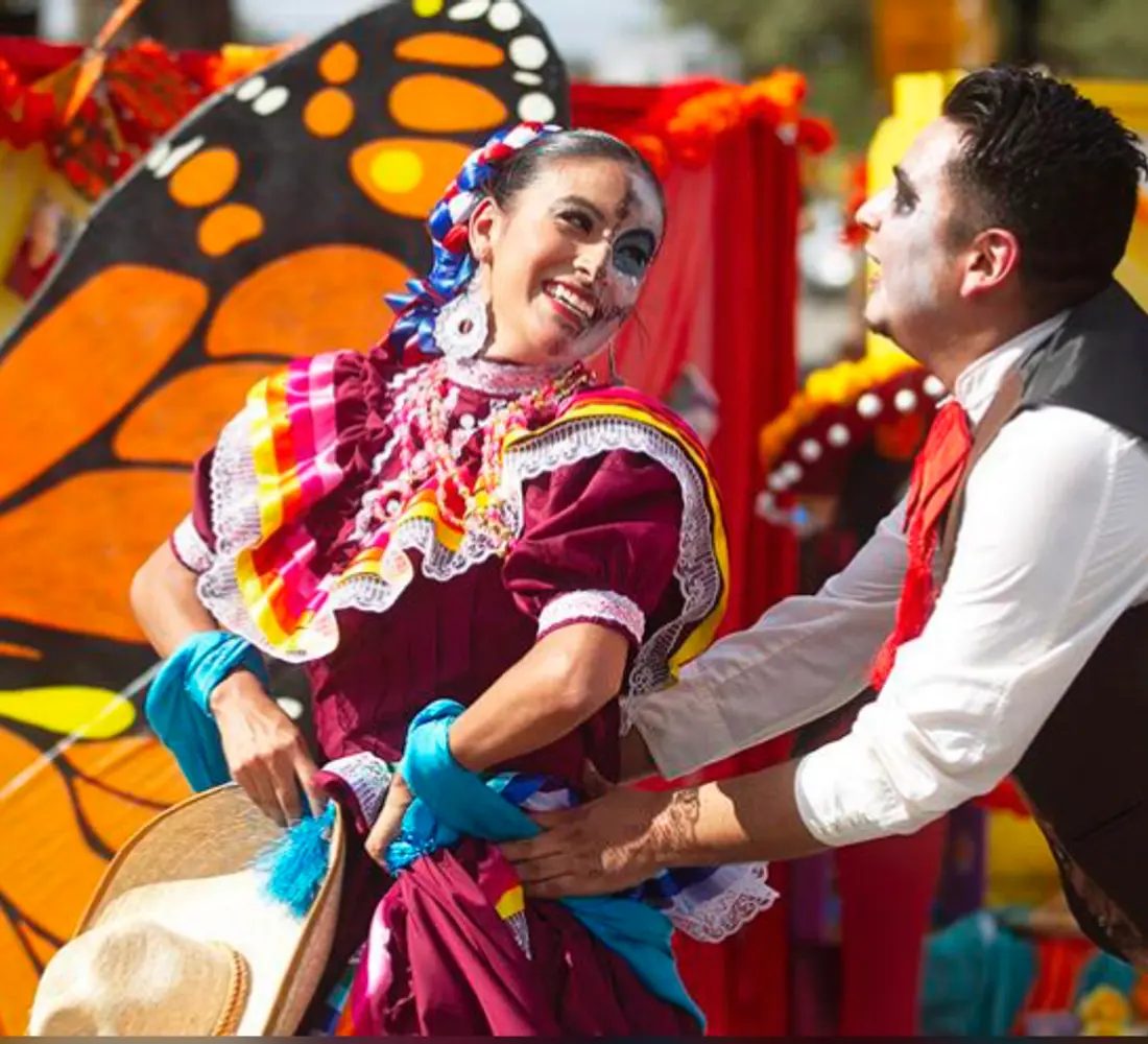 A folklórico dancer in a ruffled magenta dress and face paint dances with a partner in front of a large painted monarch butterfly.