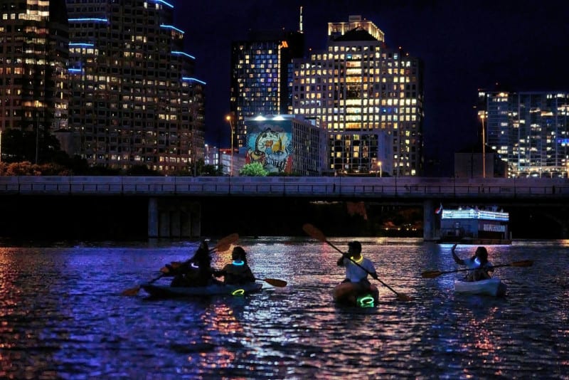 Paddlers in kayaks with glowing lights cross Lady Bird Lake at night below the lit downtown Austin skyline.