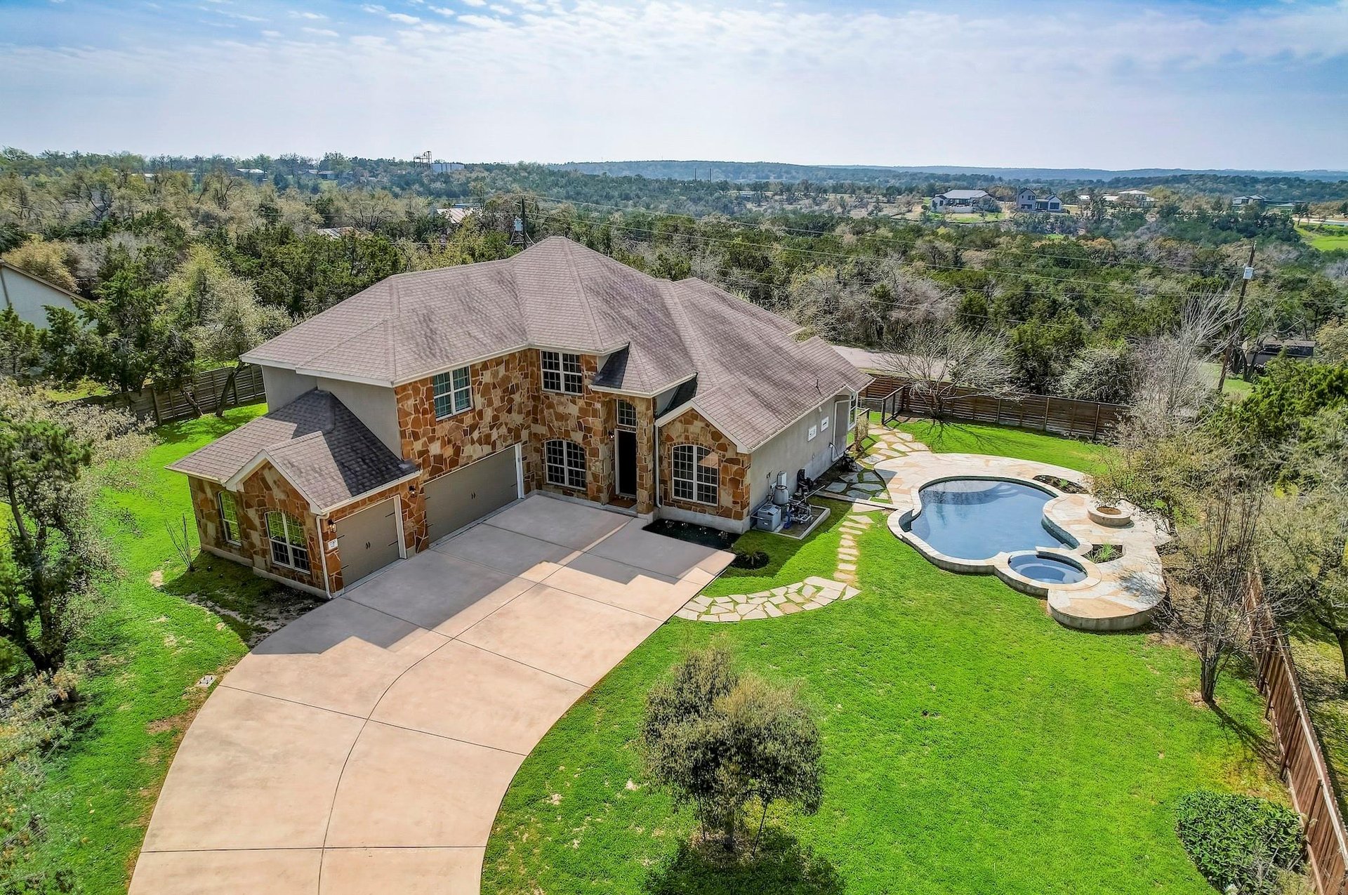 Aerial of a stone two-story home and pool on a Hill Country lot with wooded hills behind, 78736, Austin