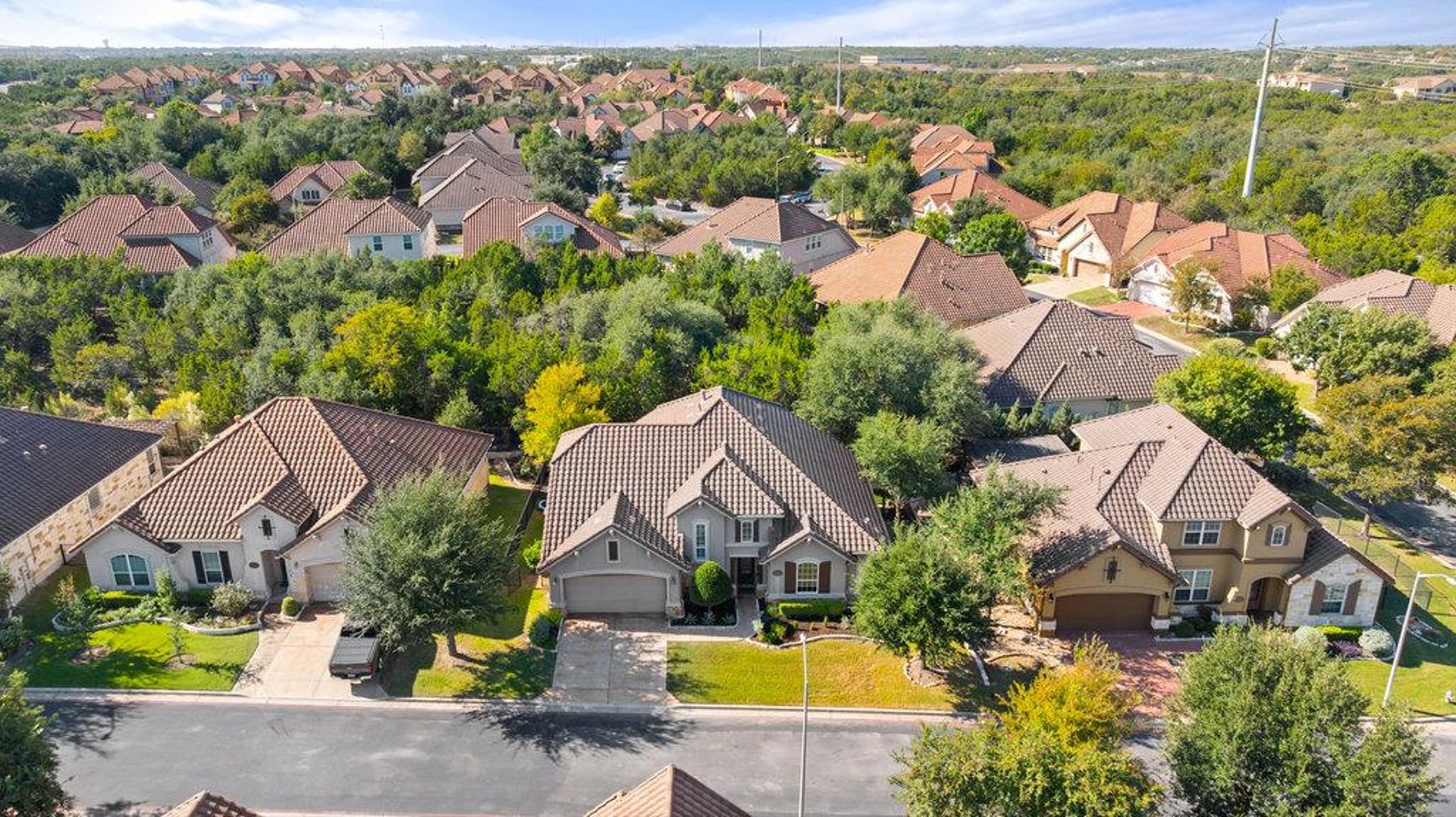 Aerial of tile-roof homes along a street with a wooded greenbelt between sections in 78726, Austin